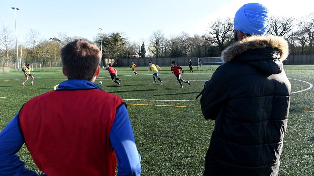 A coach and a player in the adult male grassroots game, stand side-by-side and watch the game unfold from the side of the pitch.